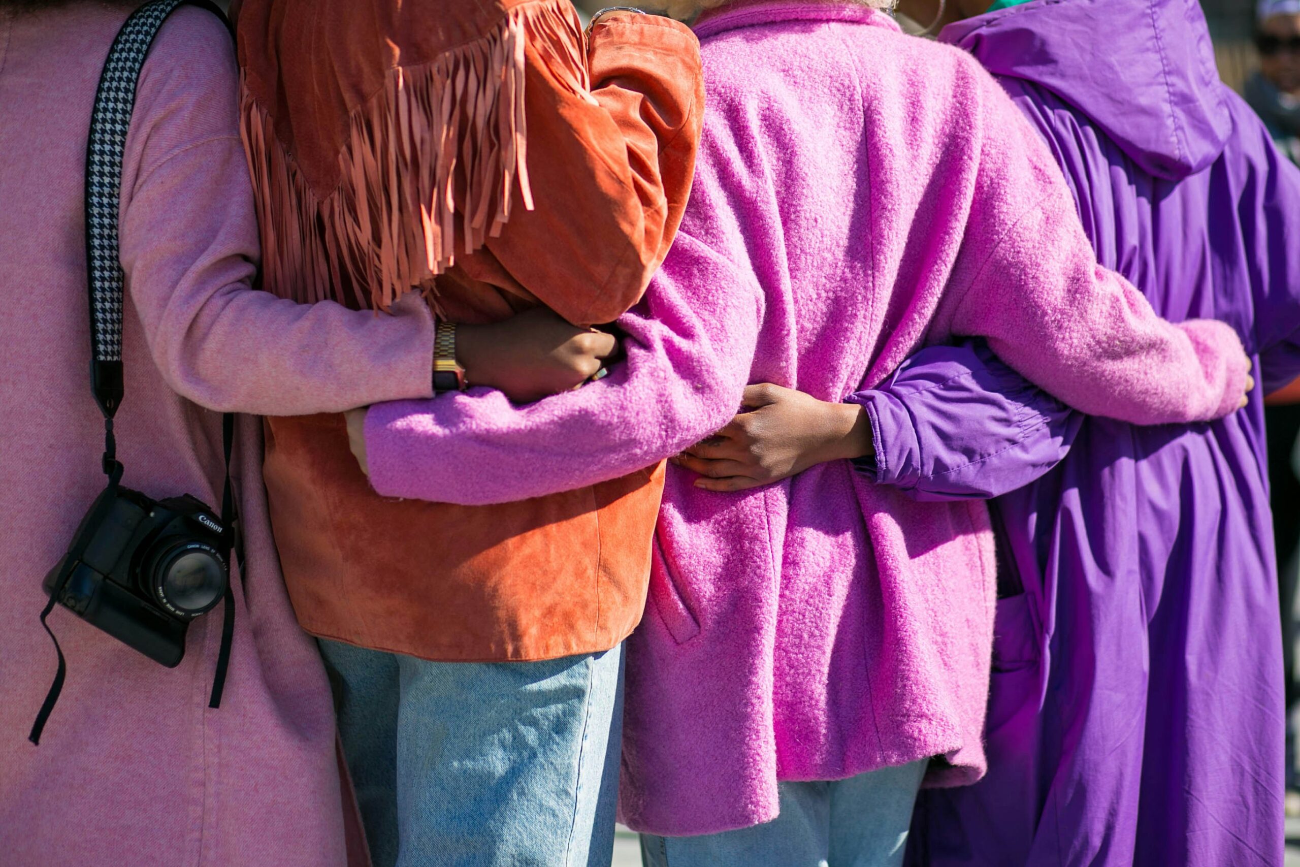 Foto på fyra personer som håller varandra om ryggen. Dom har rosa, lila och rostfärgade jackor på sig. Ansikten/huvuden syns inte.