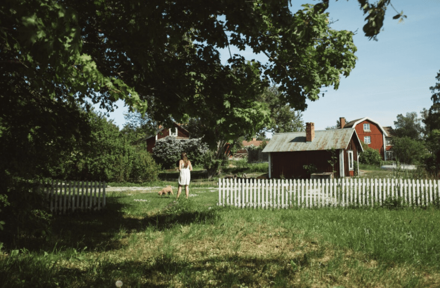 Foto av en röd stuga i grönska. Mellan stugan och ett vitt staket står en person i vit klänning med ryggen mot kameran