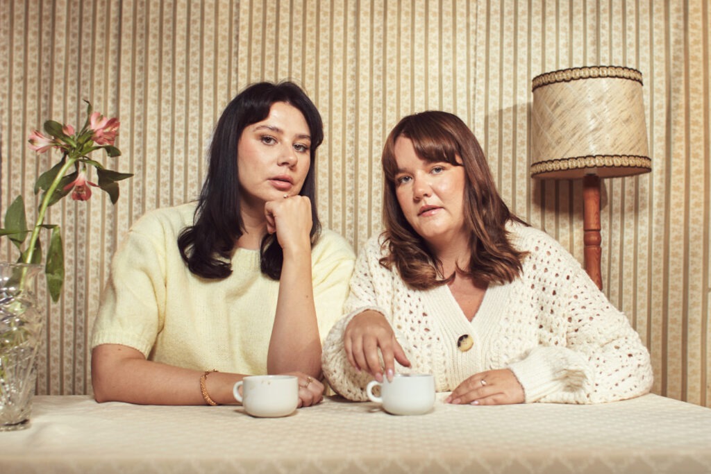 Foto på Ida Höckerstrand och Ida Hallberg (Ångestpodden) som sitter vid ett bord med kaffekoppar framför sig. Inredningen för tankarna till 50-talet.